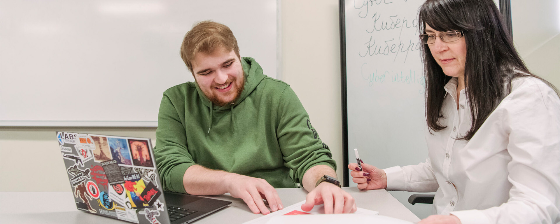 Man and woman sitting at a desk in a classroom in front of a whiteboard with Russian words, focused on the papers in front of them.