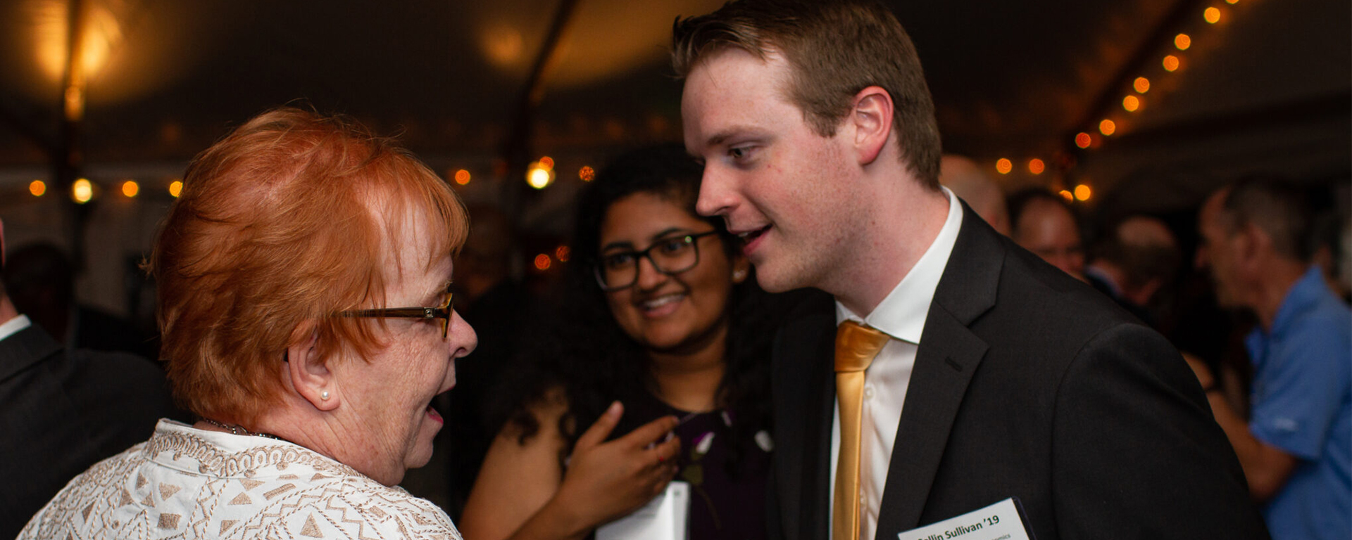 Collin Sullivan ’19, Digital Credential leader, wearing a black suit and gold tie, shakes hands with an older woman in a white patterned jacket at an event. The interaction is warm and professional, with Collin holding a yellow program booklet. In the background, a woman with curly hair and glasses smiles, adding to the lively atmosphere of the gathering