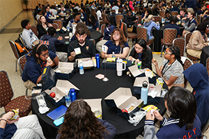 A group of young people seated at a round table eat lunch at conference event with other groups seated in background.